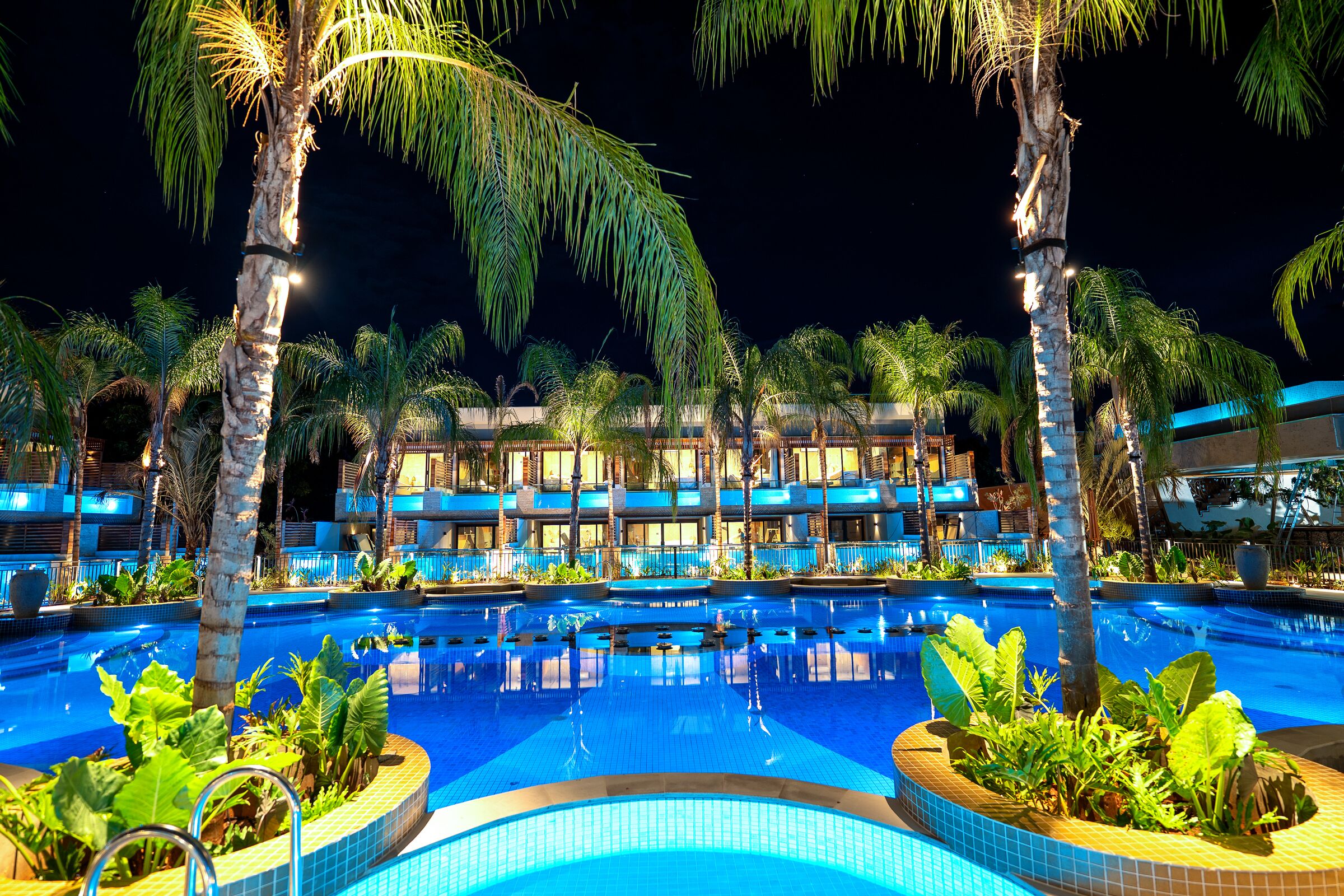 A swimming pool surrounded by palm trees and a building with multiple balconies.