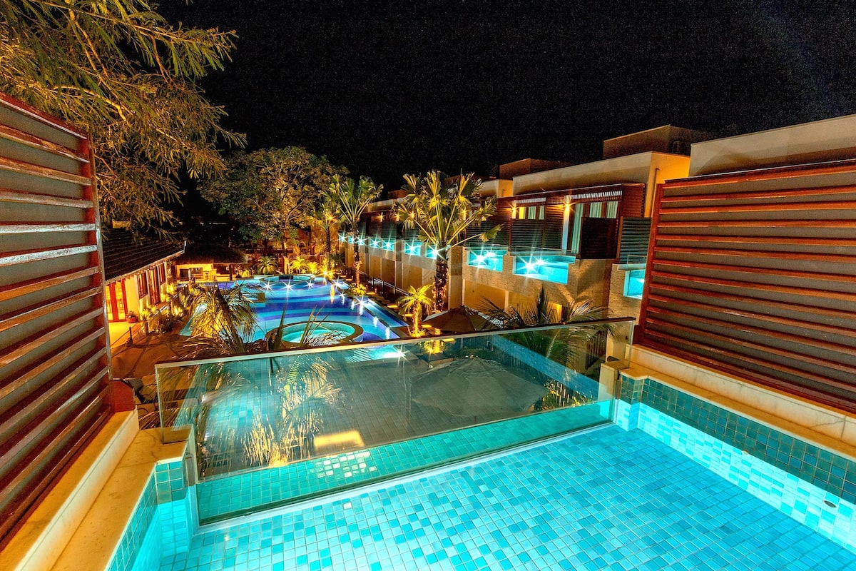 Vista nocturna de un hotel con piscina, palmeras y arquitectura moderna.
