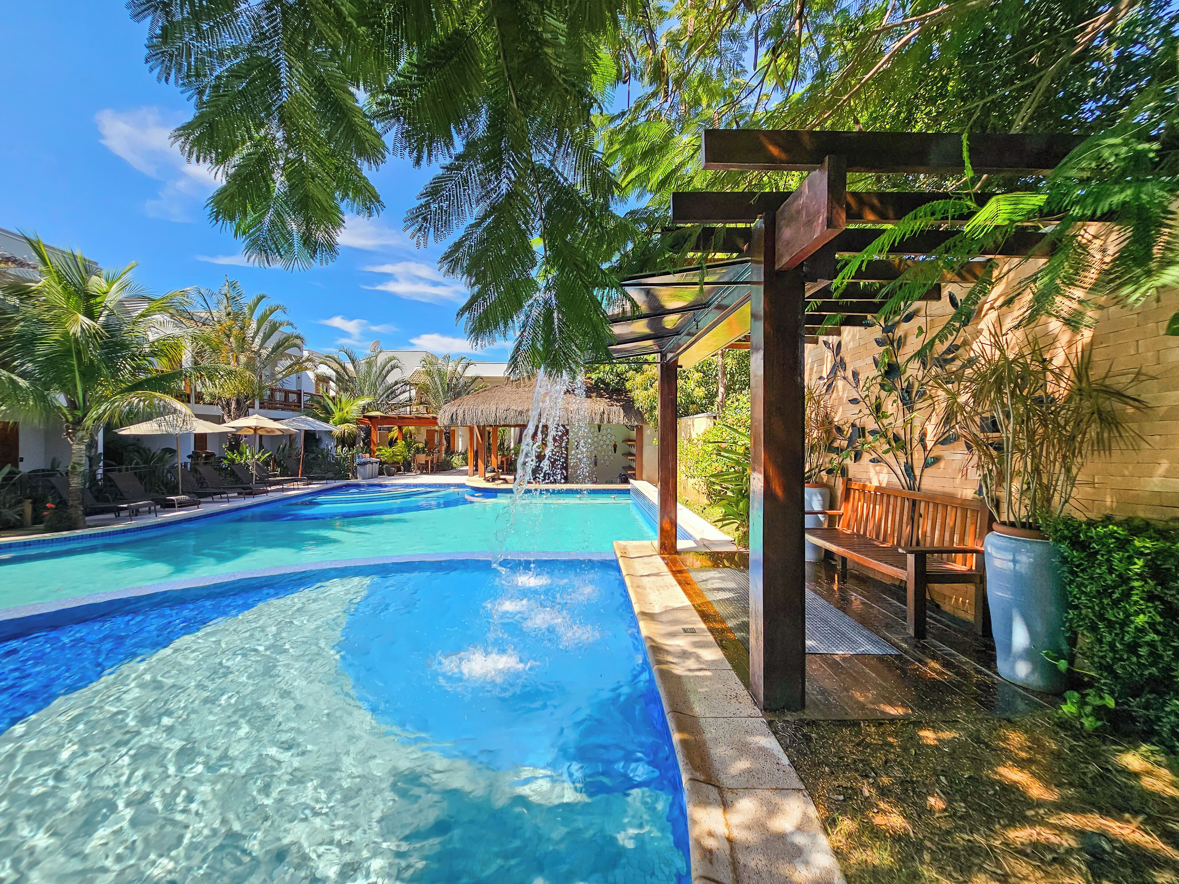 A clear blue swimming pool with a wooden deck and lounge chairs, surrounded by tropical plants and a thatched-roof structure.