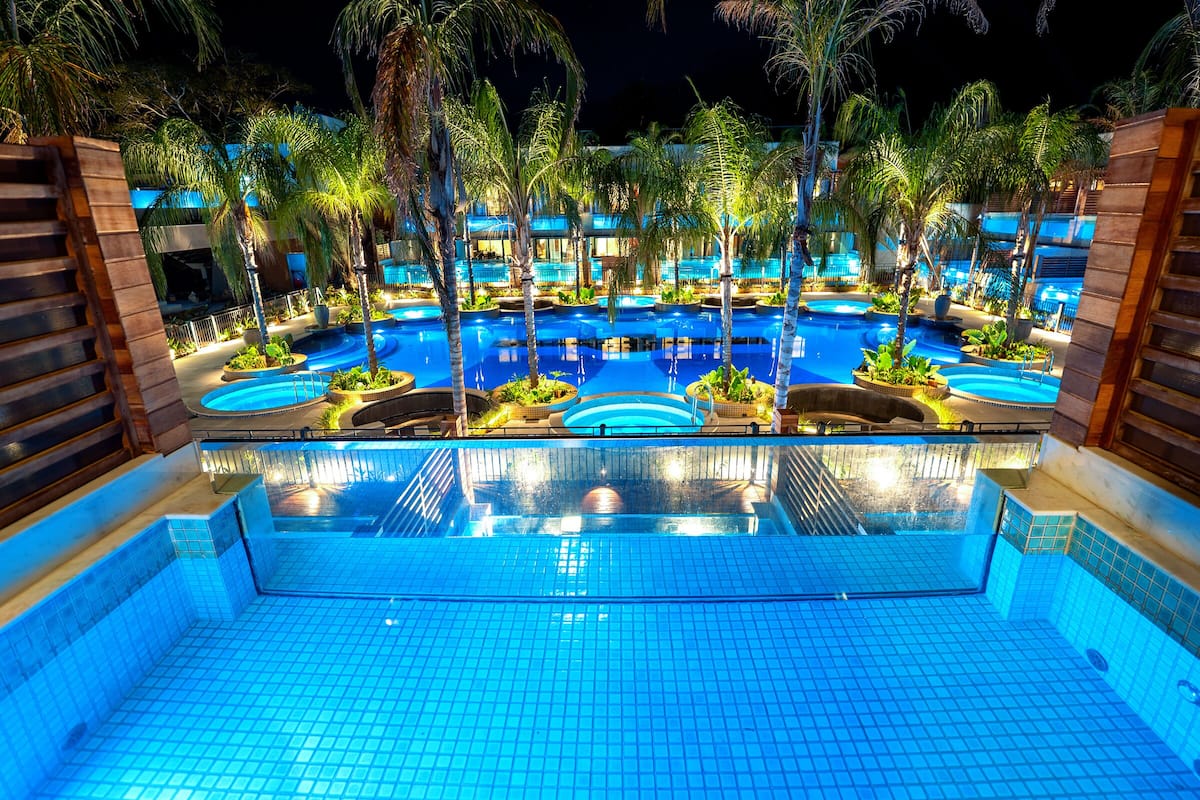 Piscina con varios niveles y palmeras circundantes, rodeada de edificios.