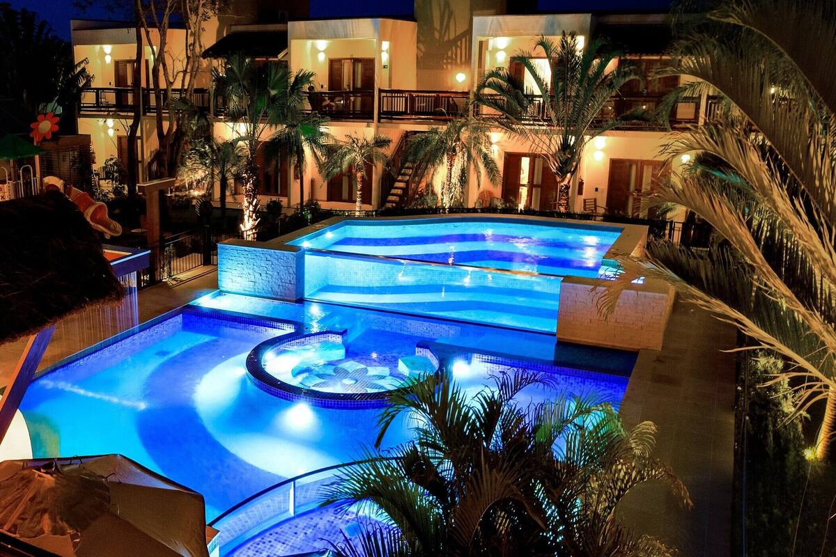 Un hotel de varios niveles con una amplia piscina iluminada rodeada de palmeras.