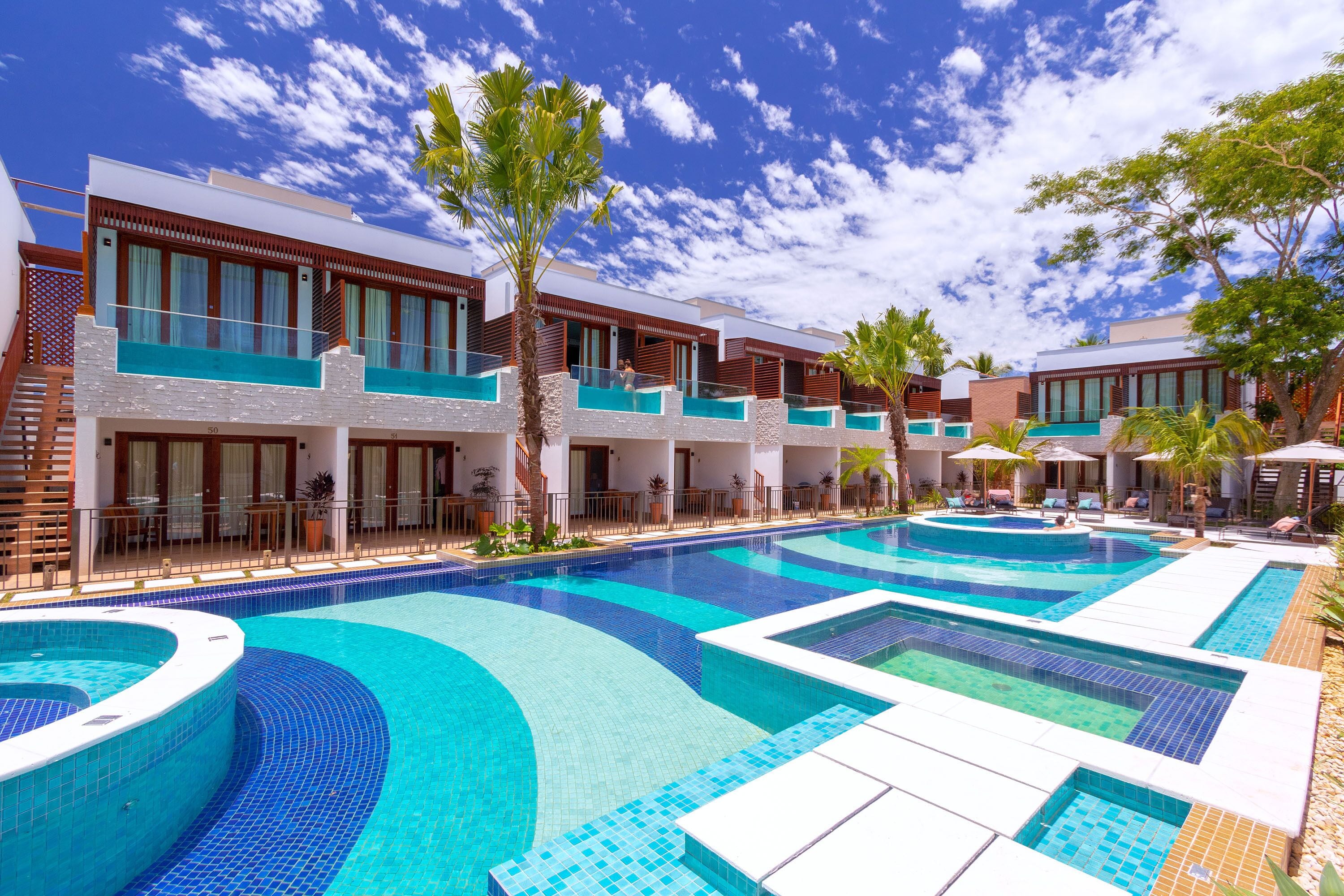 A modern hotel with multiple swimming pools, a covered outdoor area, and a clear blue sky.