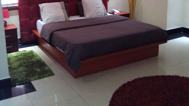 A bedroom with a large bed, a nightstand, a red headboard, a window with blinds, and a green rug.