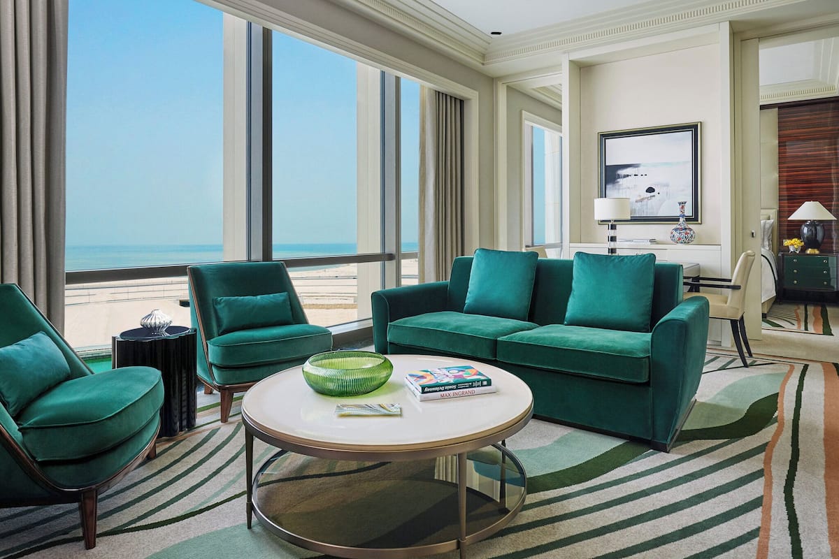A modern living room with a large window offering a view of the beach. There is a green sofa, a green armchair, and a round coffee table with a book and a bowl on it.