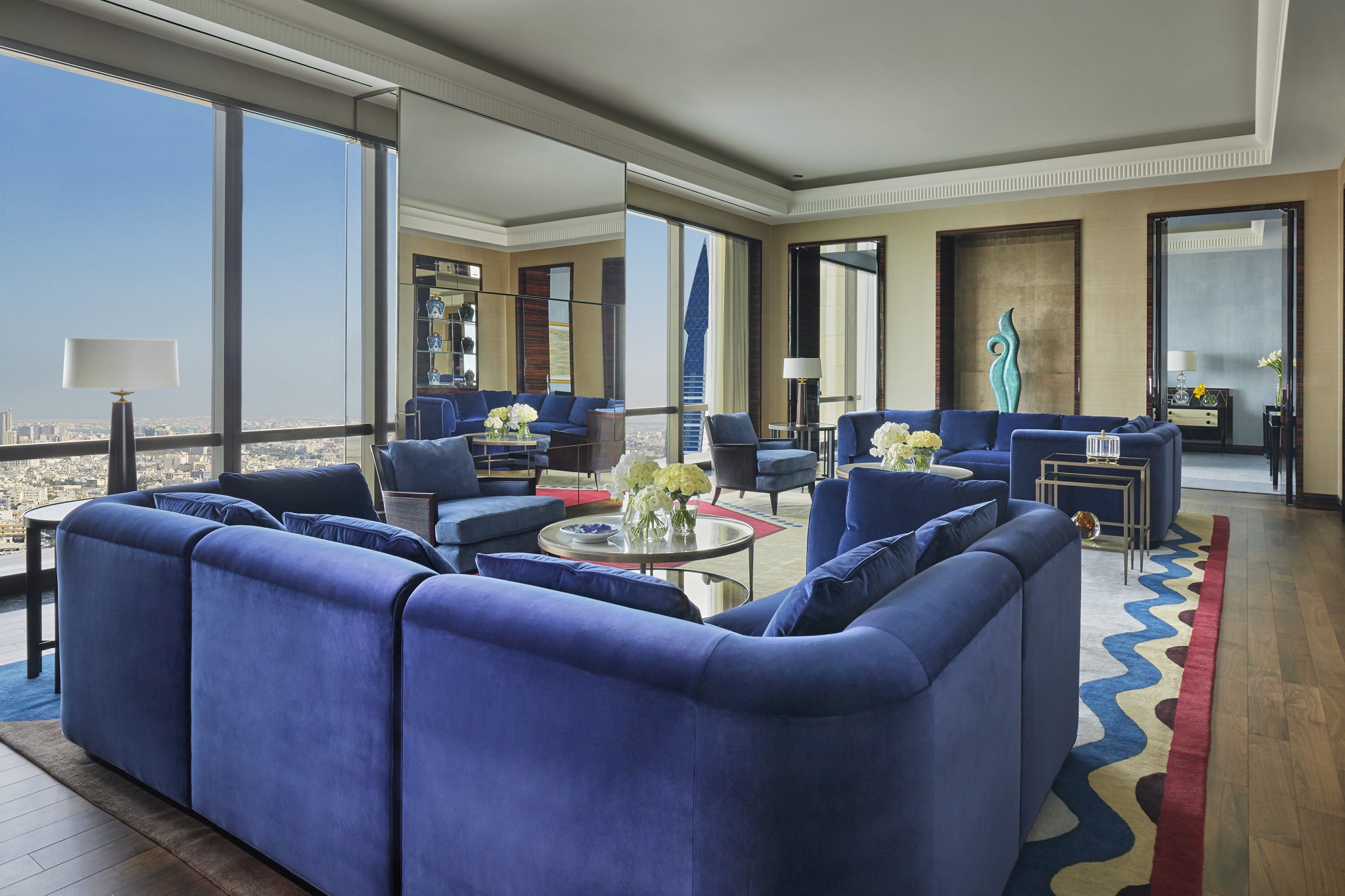 A modern hotel room with a large window offering a city view, a seating area with blue sofas, and a small table with flowers.