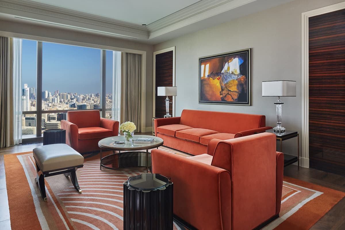 A modern hotel room with a large window offering a city view, a sofa, a chair, a small table with a vase, and a painting on the wall.