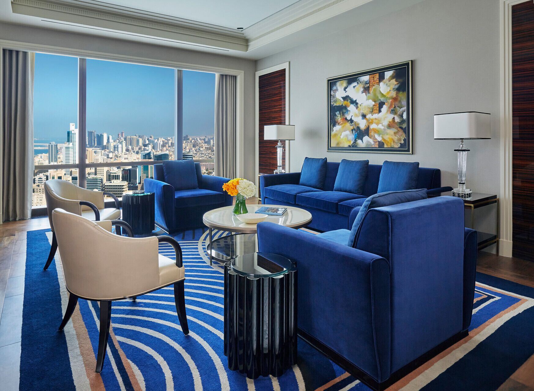 Habitación de hotel moderna con vistas a la ciudad, un sofá azul, una mesita de centro redonda y un cuadro abstracto grande.