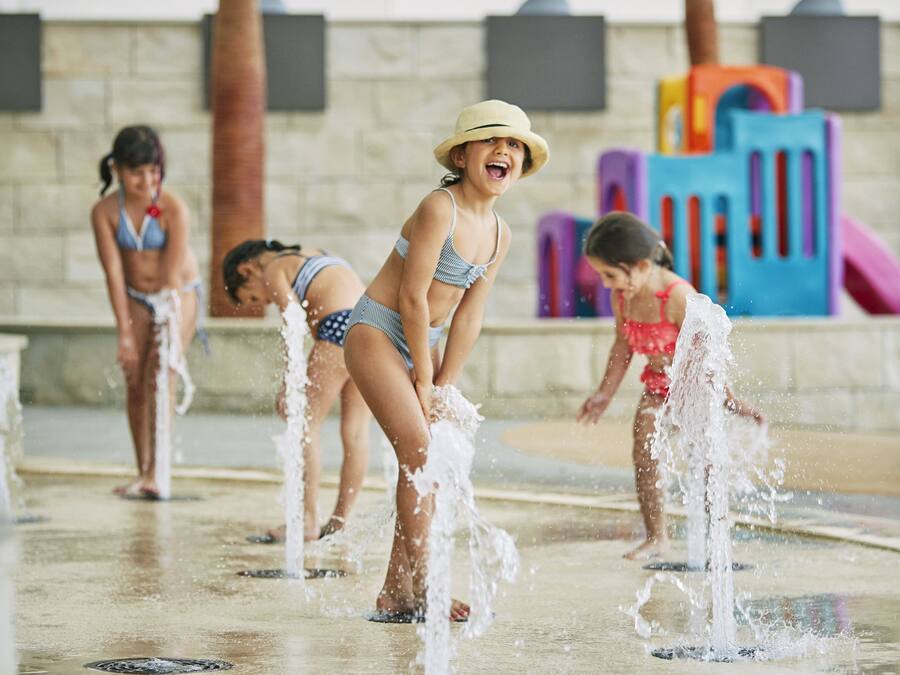 A water play area with multiple water jets and colorful play structures in the background.