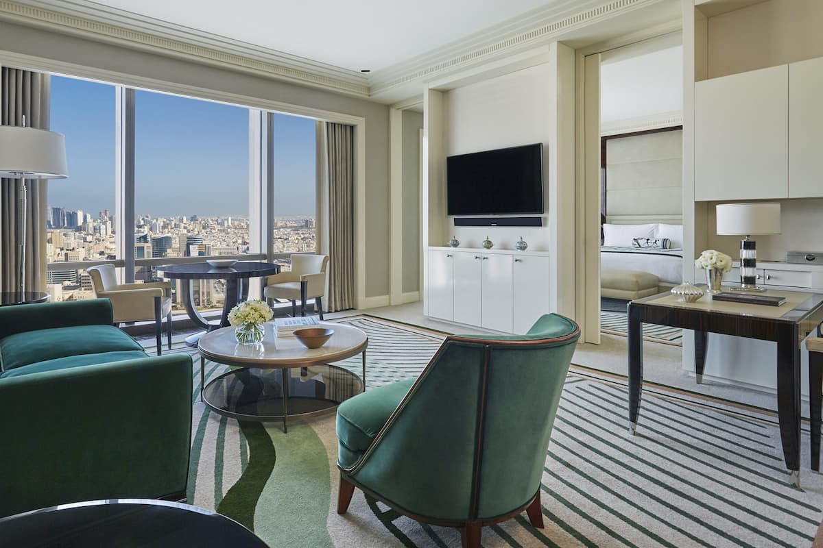 A modern hotel room with a city view, a dining area, and a living room with green armchairs and a round coffee table.