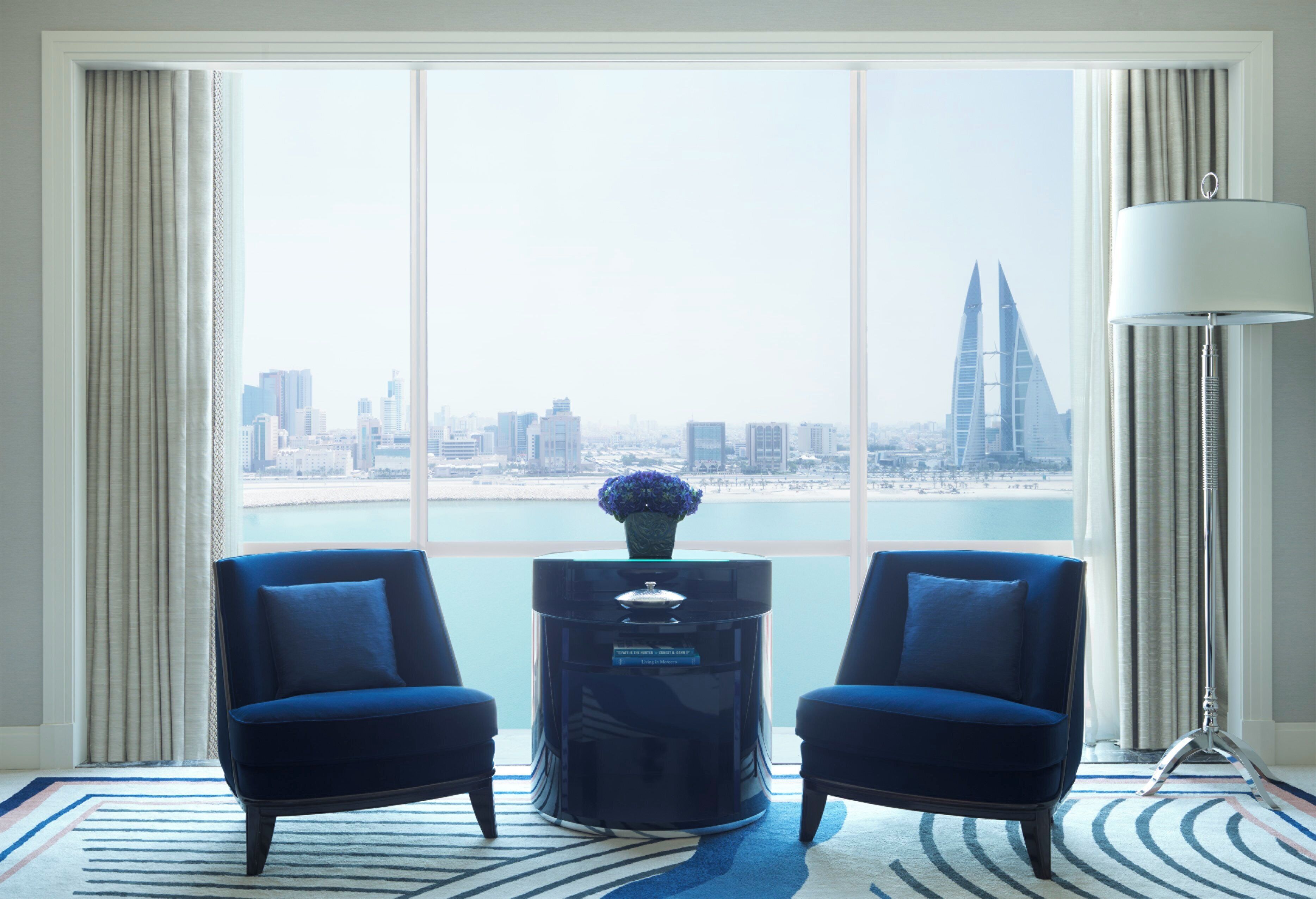 A room with two blue armchairs, a black side table with a vase, a floor lamp, and a large window with a city view.