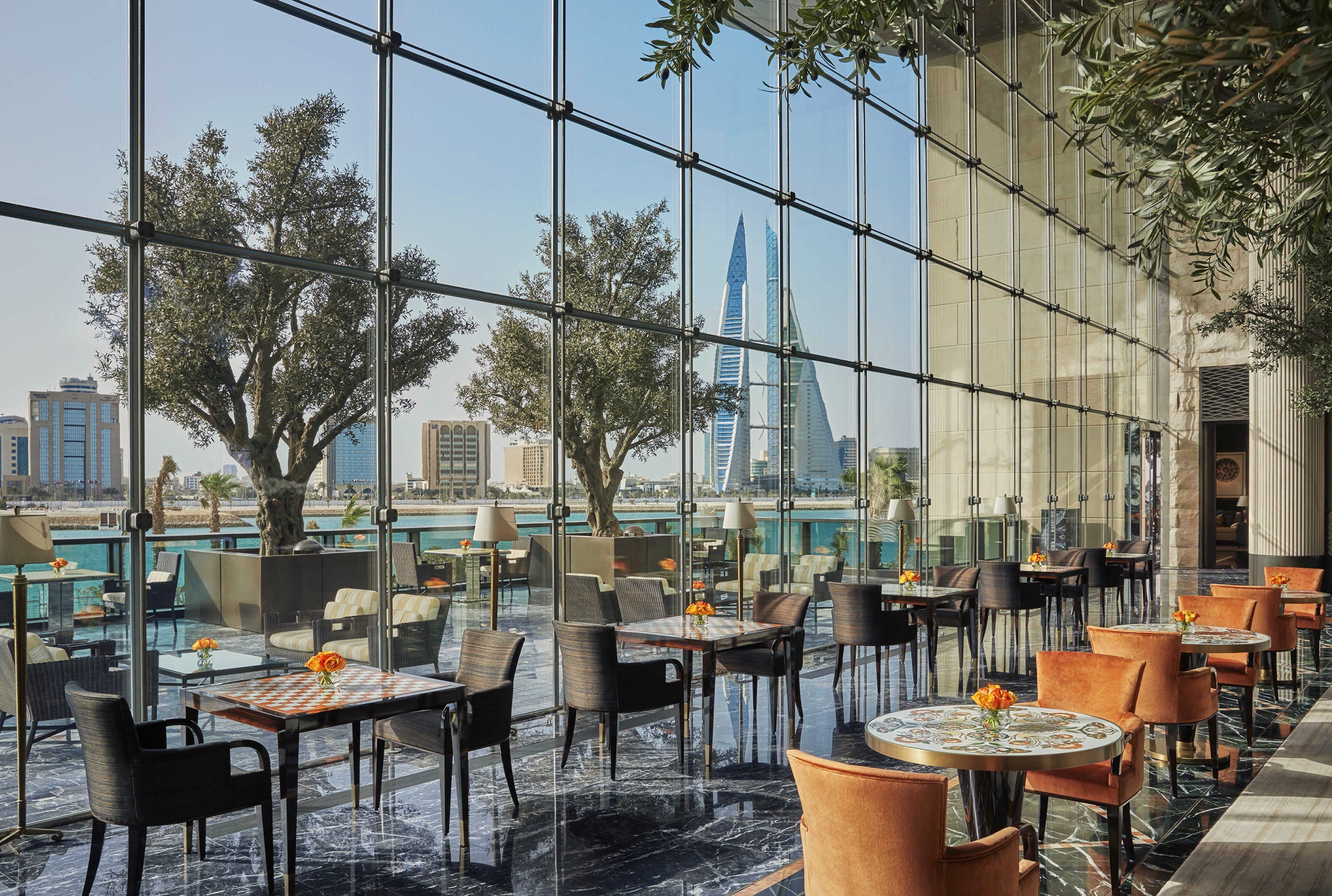 Un comedor interior moderno con mesas y sillas, ventanas amplias y vistas a un edificio alto y palmeras.