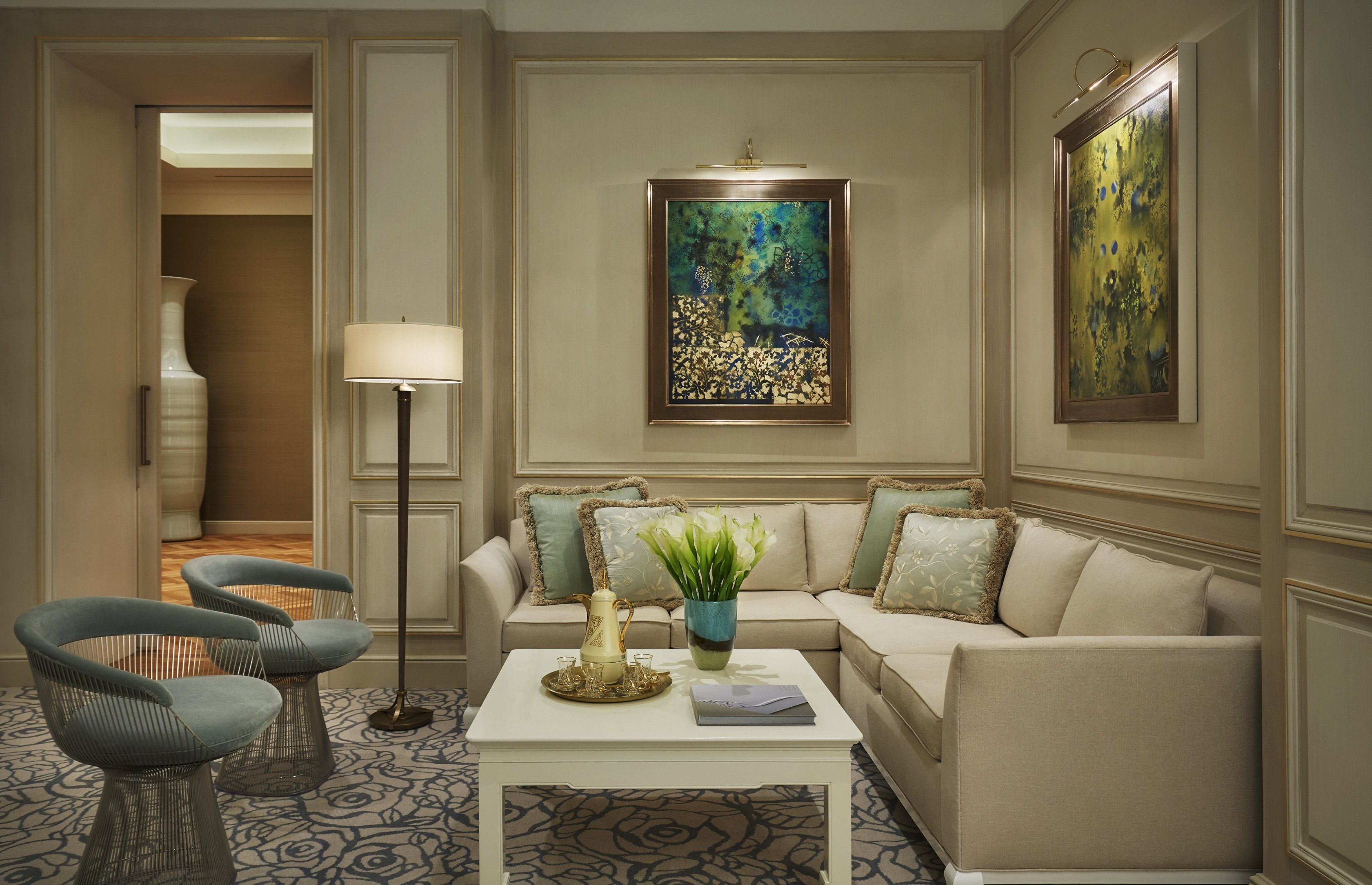 Una sala de estar con un sofá color beige, dos sillones azules, una mesa de centro con objetos decorativos, una lámpara de pie y un cuadro en la pared.