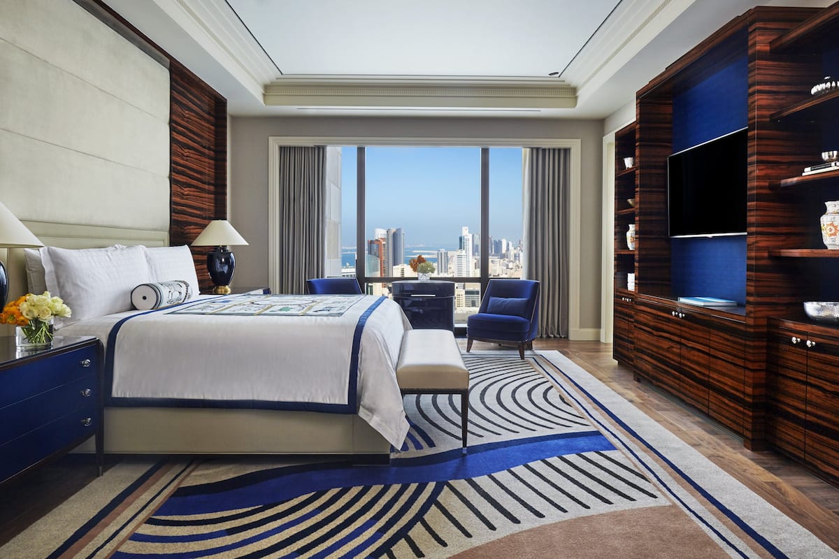 A hotel room with a large bed, a desk, a chair, a TV, and a view of the city.