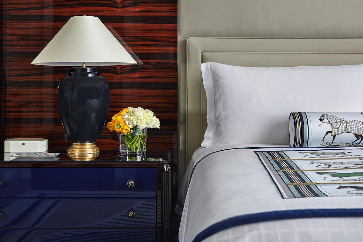 A neatly arranged hotel room with a bed, a bedside table with a lamp, a vase with flowers, and a nightstand with drawers.