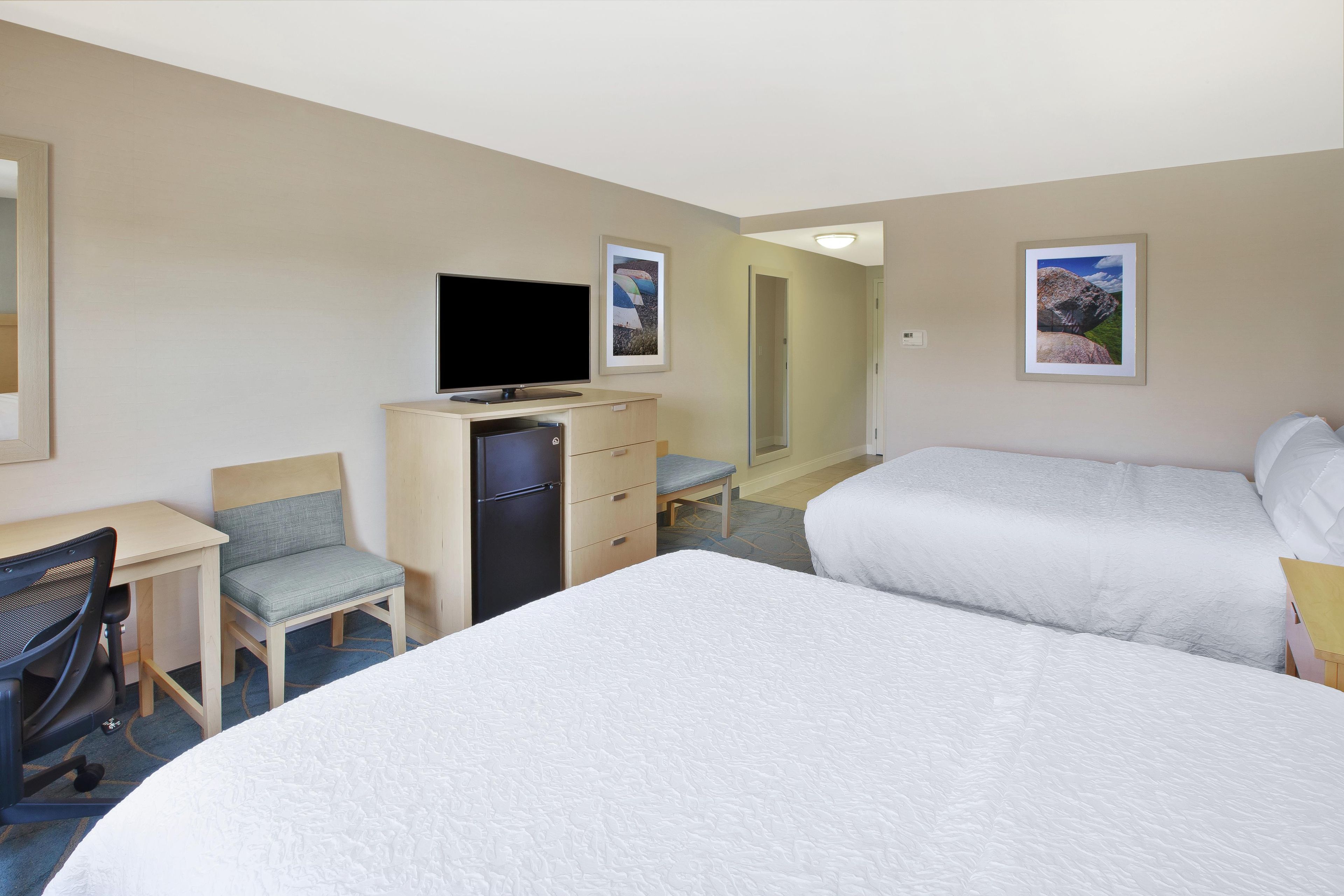 A hotel room with two beds, a desk, a chair, a television, and artwork on the walls.
