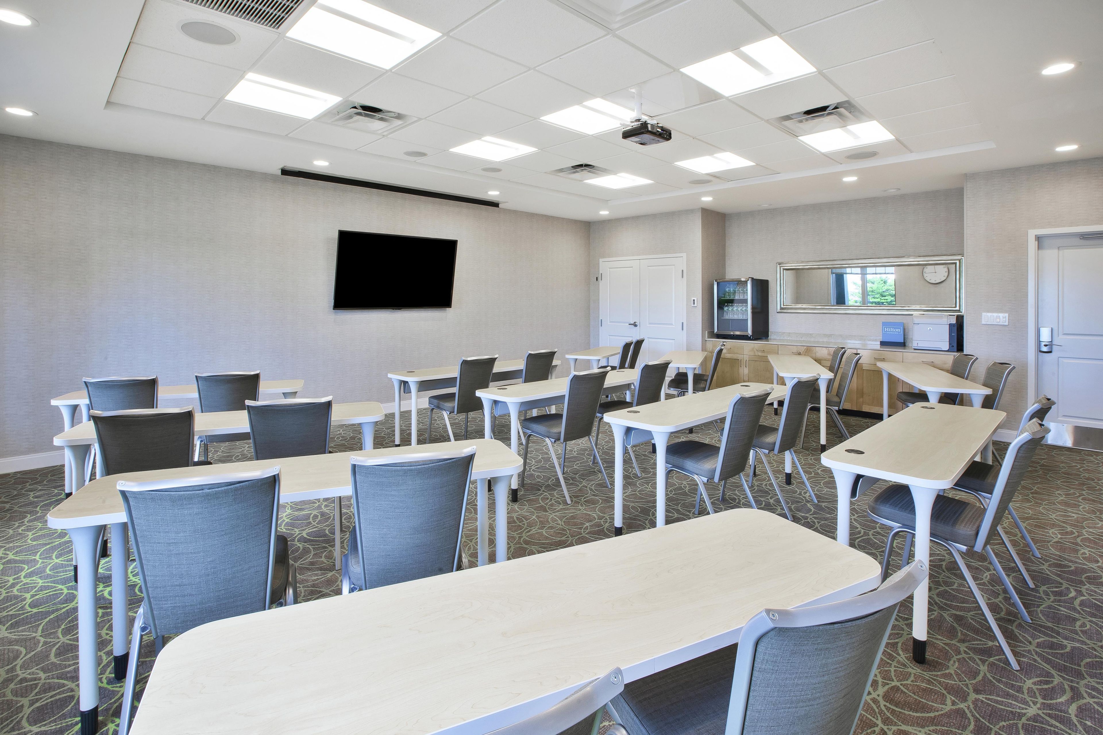 A conference room with tables and chairs, a flat-screen TV, a clock, and a minibar.