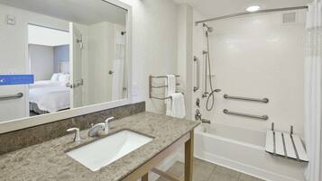A modern bathroom with a large sink, a shower with grab bars, and a mirror reflecting a bedroom.