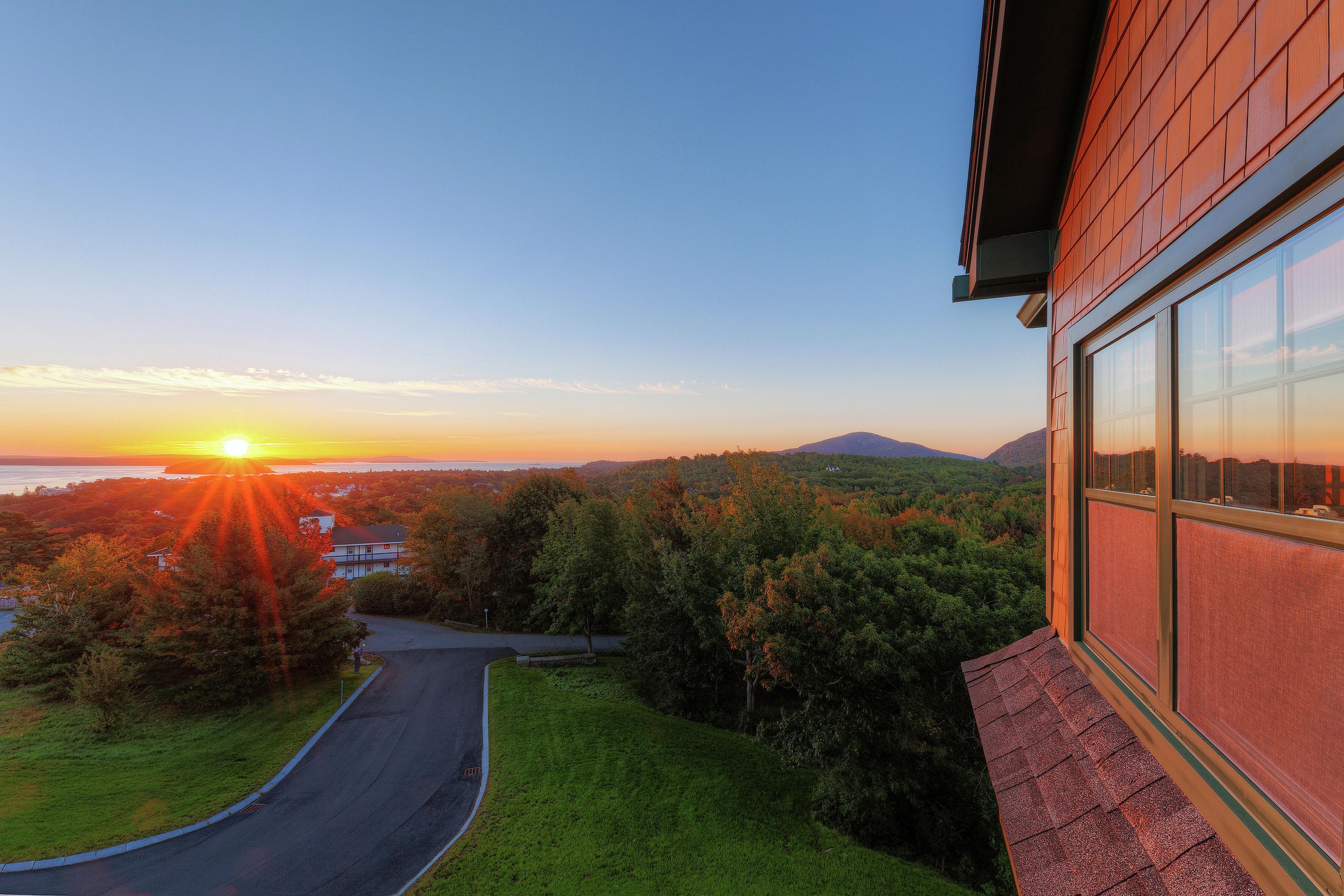 A residential property with a driveway leading to a house, surrounded by trees and a view of a sunset over a body of water.