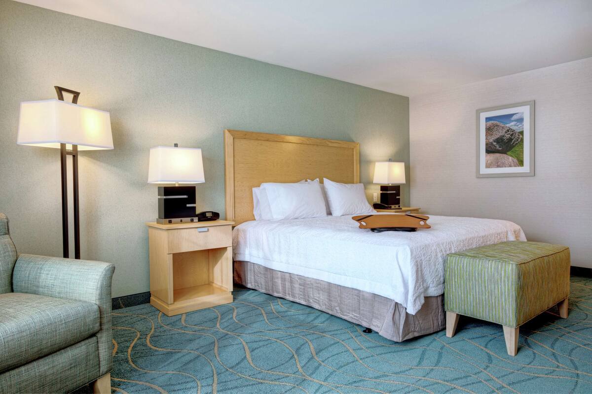 A hotel room with a bed, two bedside lamps, a small wooden nightstand, a green upholstered chair, and a matching ottoman.