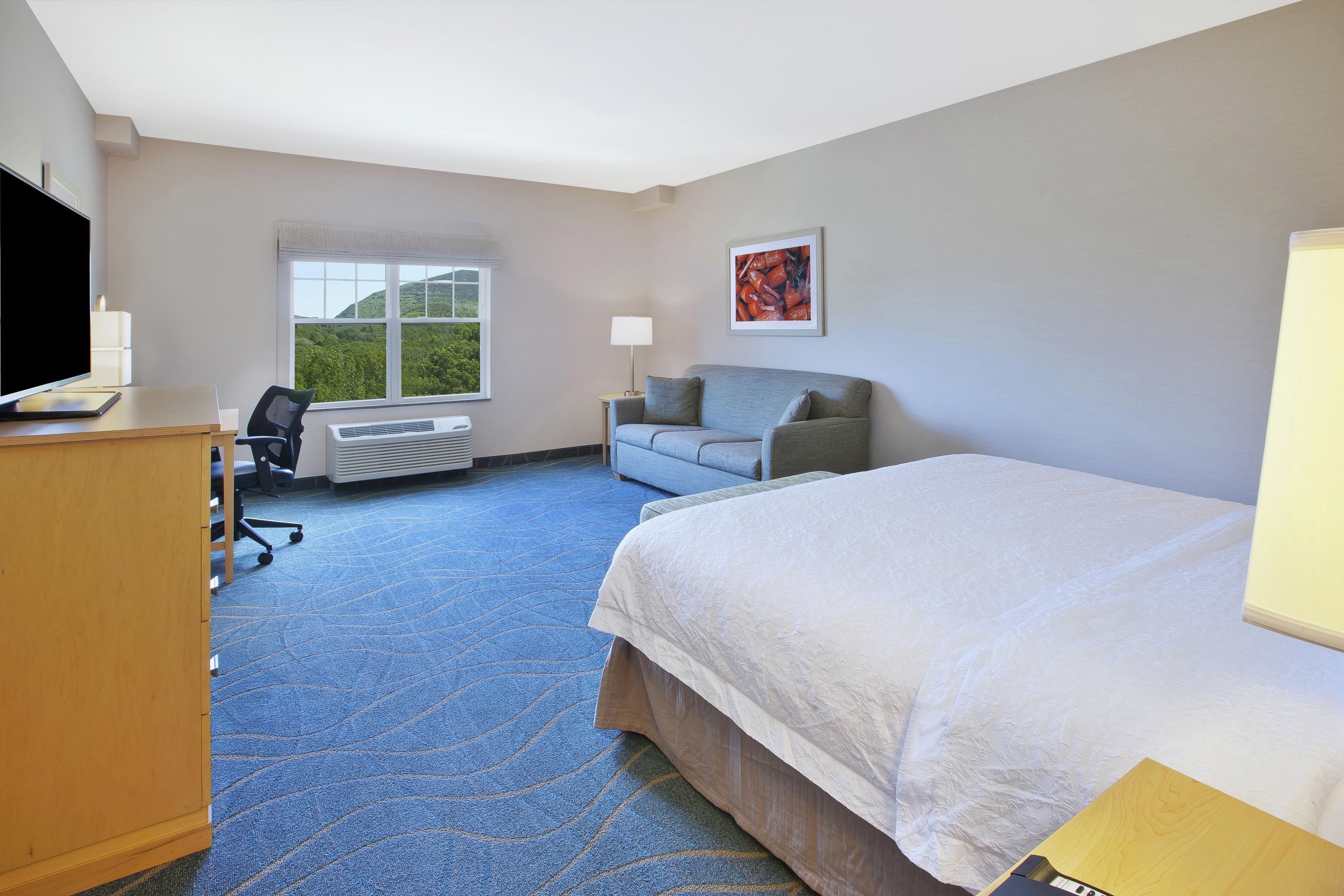 A hotel room with a bed, a desk with a computer, a chair, a window with a view of greenery, and a painting on the wall.