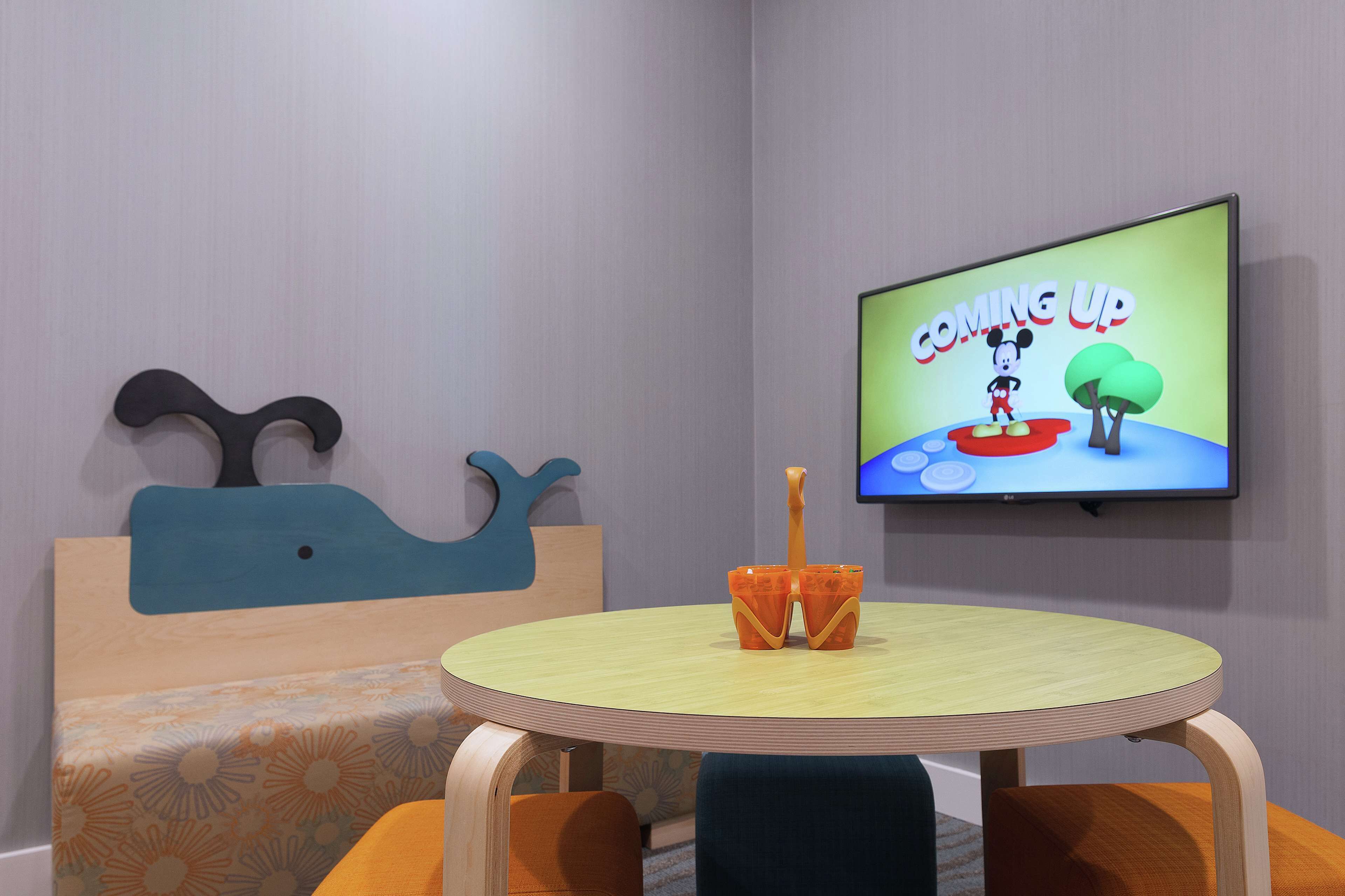 A room with a round wooden table, a television displaying 'COMING UP' with a cartoon character, a blue whale-shaped headboard, and a patterned bedspread.