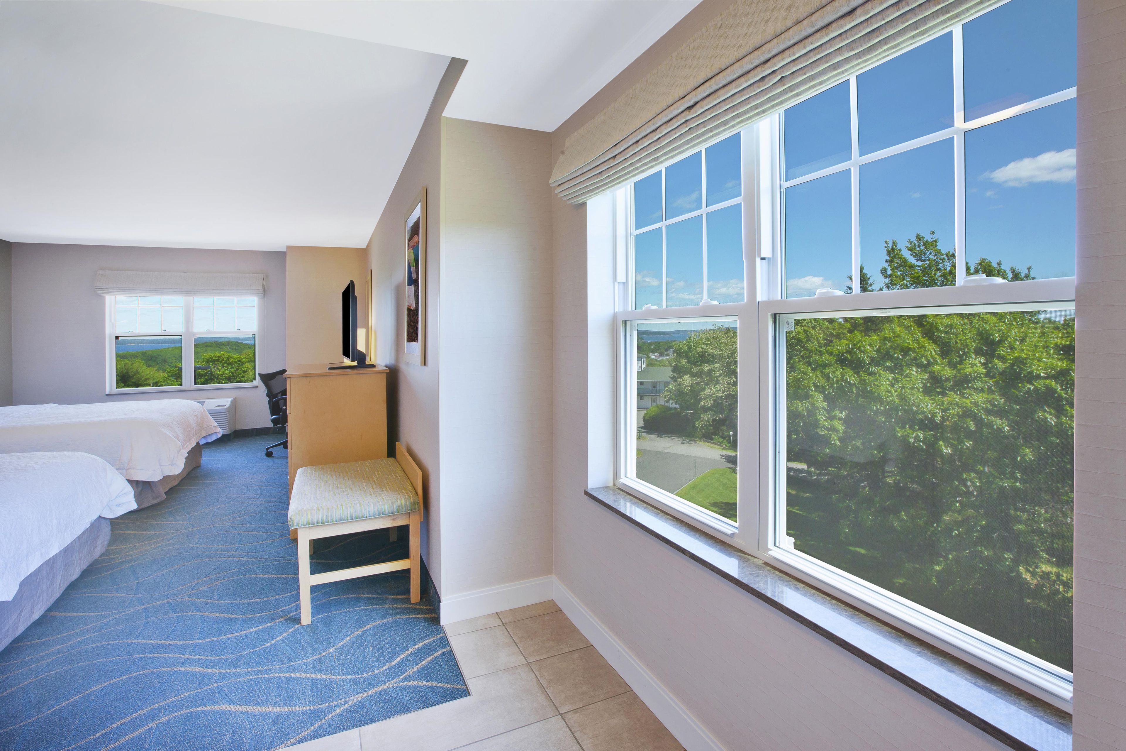 Une chambre d’hôtel avec deux lits, un bureau, une chaise et une grande fenêtre donnant sur un paysage verdoyant.