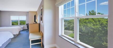 Une chambre d’hôtel avec deux lits, un bureau, une chaise et une grande fenêtre donnant sur un paysage verdoyant.
