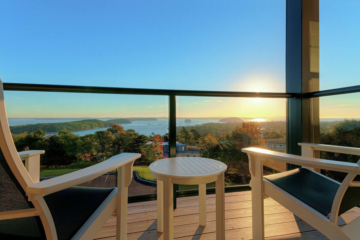 A balcony with two chairs and a small table, overlooking a lake and forested area.
