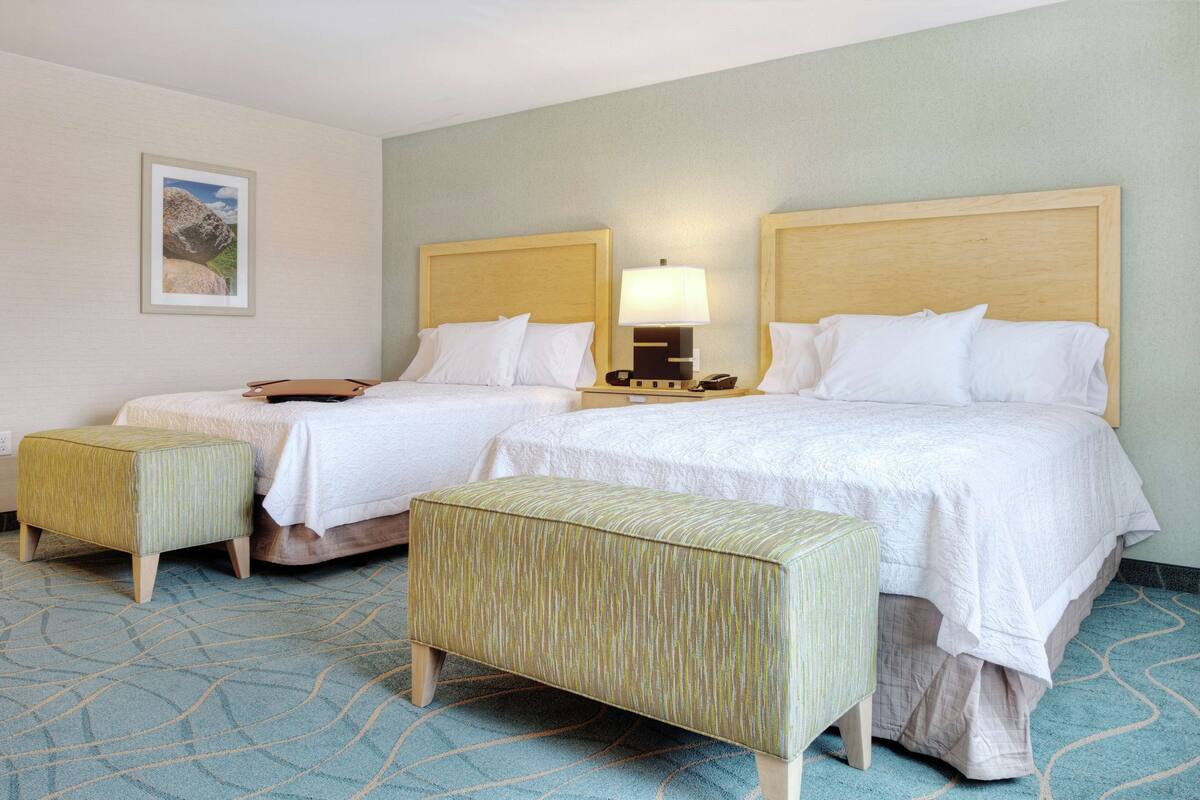 A hotel room with two beds, a nightstand with a lamp, and a small stool.