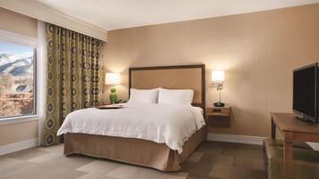 Habitación de hotel con una cama grande, dos lámparas de noche, un televisor sobre una mesa de madera y vistas a montañas a través de la ventana.