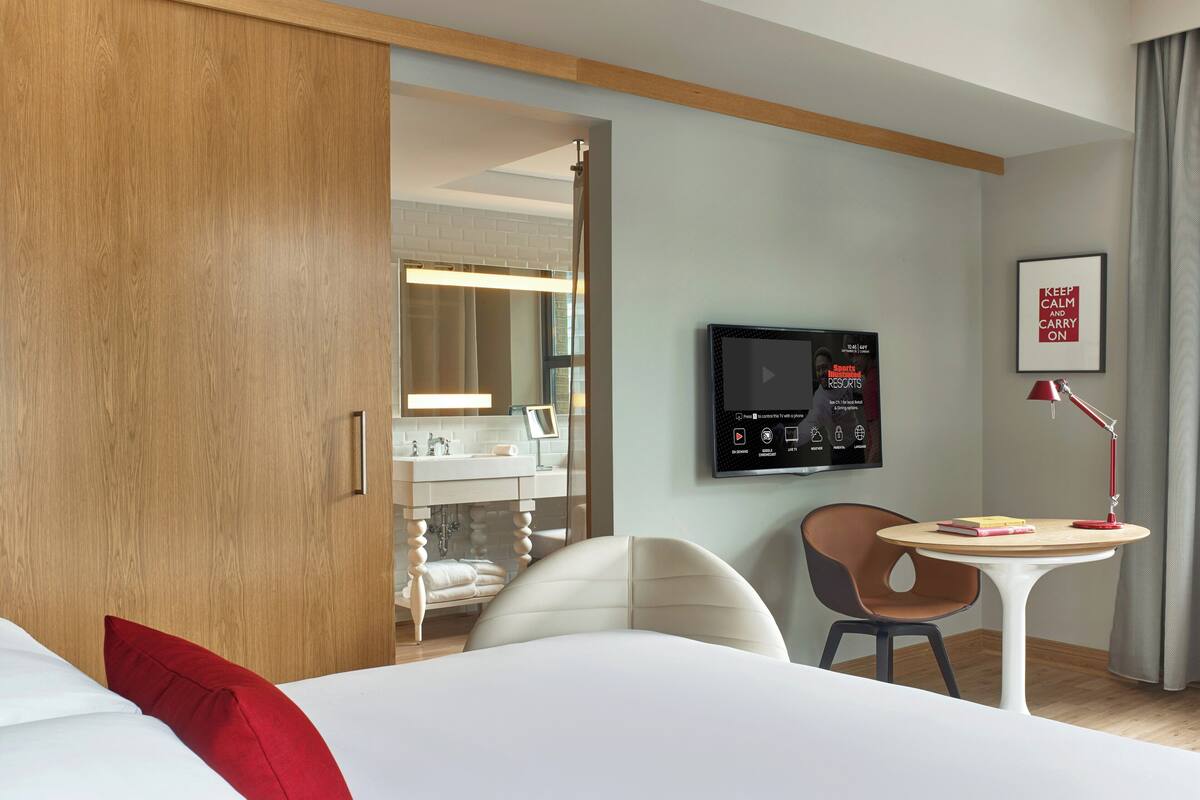 A hotel room with a bed, a desk, a chair, a TV, and a bathroom visible through an open door.