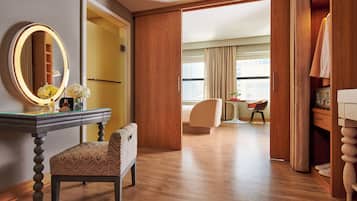 Quarto de hotel com piso de madeira, uma escrivaninha com espelho, uma cadeira, um guarda-roupa e vista da cidade por grandes janelas.