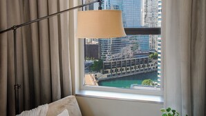 Ein Hotelzimmer mit Blick auf eine Stadtlandschaft durch ein großes Fenster, eine Stehlampe, eine Couch mit Kissen und ein kleiner Tisch mit einer Pflanze.