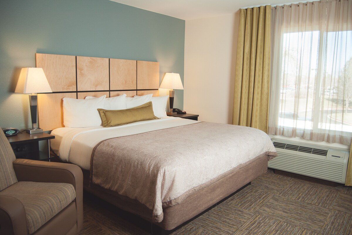 A hotel room with a bed, two bedside tables, a chair, a window with curtains, and an air conditioning unit.