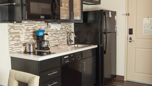 A compact kitchen with dark cabinets, a microwave, a refrigerator, and a coffee maker. - Candlewood Suites Fort Collins by IHG (Fort Collins)