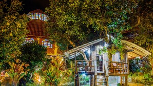 A well-lit garden with a wooden structure, surrounded by lush greenery and a large tree. - Hosteria Jardines de Chamana (Ulba)
