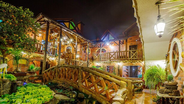 A rustic courtyard with a wooden bridge, a small pond, and a brick building with multiple balconies and outdoor lighting. - Hosteria Jardines de Chamana (Ulba)