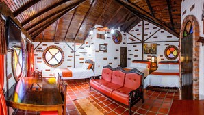 A cozy, rustic room with a high ceiling, wooden beams, and a tiled floor. There are two beds with white bedding and red accents, a red leather sofa, and a wooden table with chairs. - Hosteria Jardines de Chamana (Ulba)