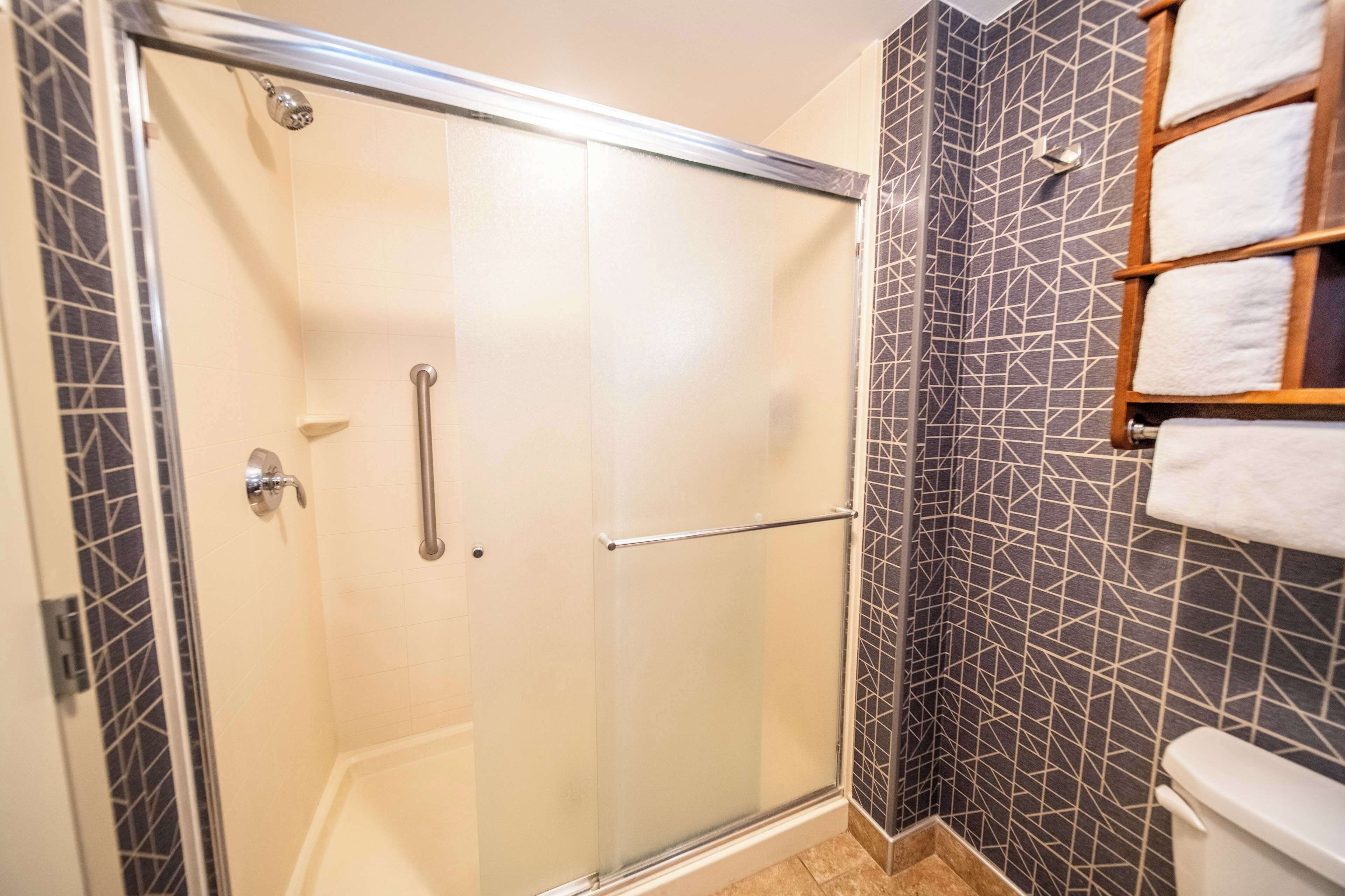 A shower with a glass door, a handrail, and a tiled wall with a geometric pattern.
