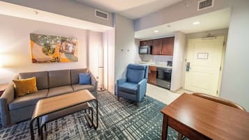 Una sala de estar moderna con sofá, sillón y mesa de centro, un área de cocina con microondas y lavavajillas, y una puerta con un letrero.