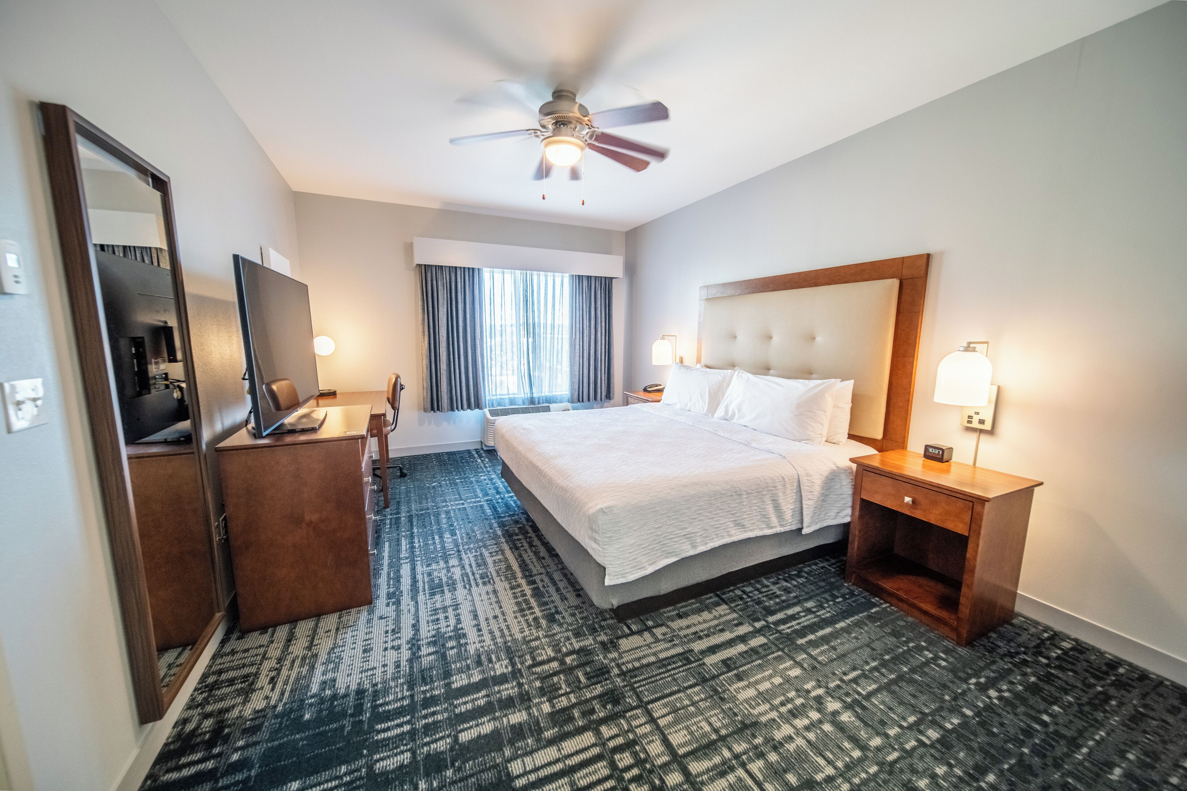 Une chambre d’hôtel équipée d’un lit, d’un bureau, d’un miroir, d’un ventilateur de plafond et d’une fenêtre avec des rideaux.