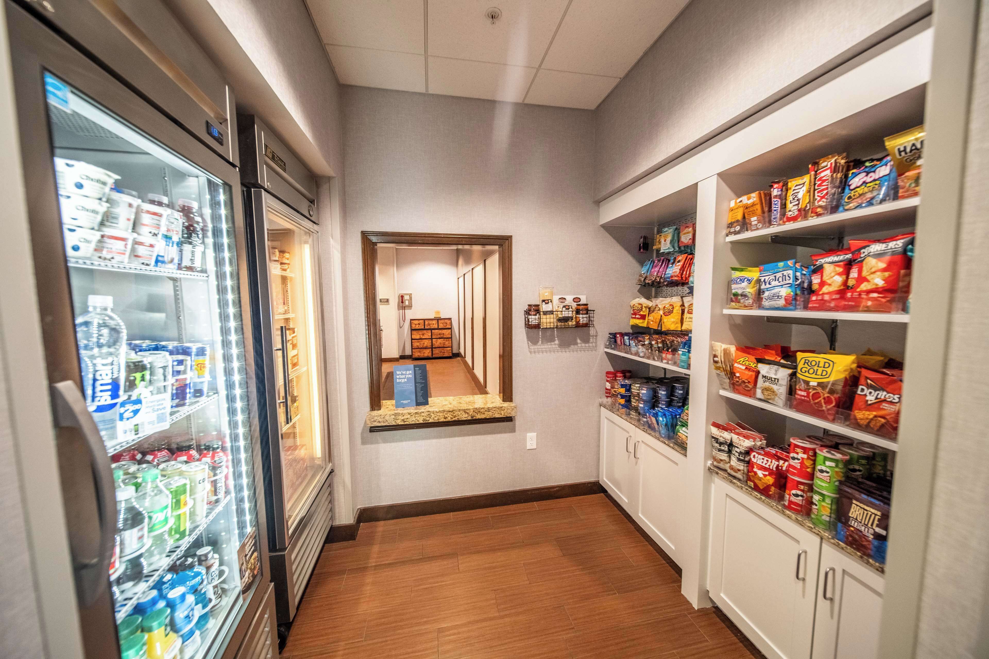 A narrow hallway with a refrigerator stocked with beverages and snacks, leading to a room with a wooden floor and a view through a doorway.