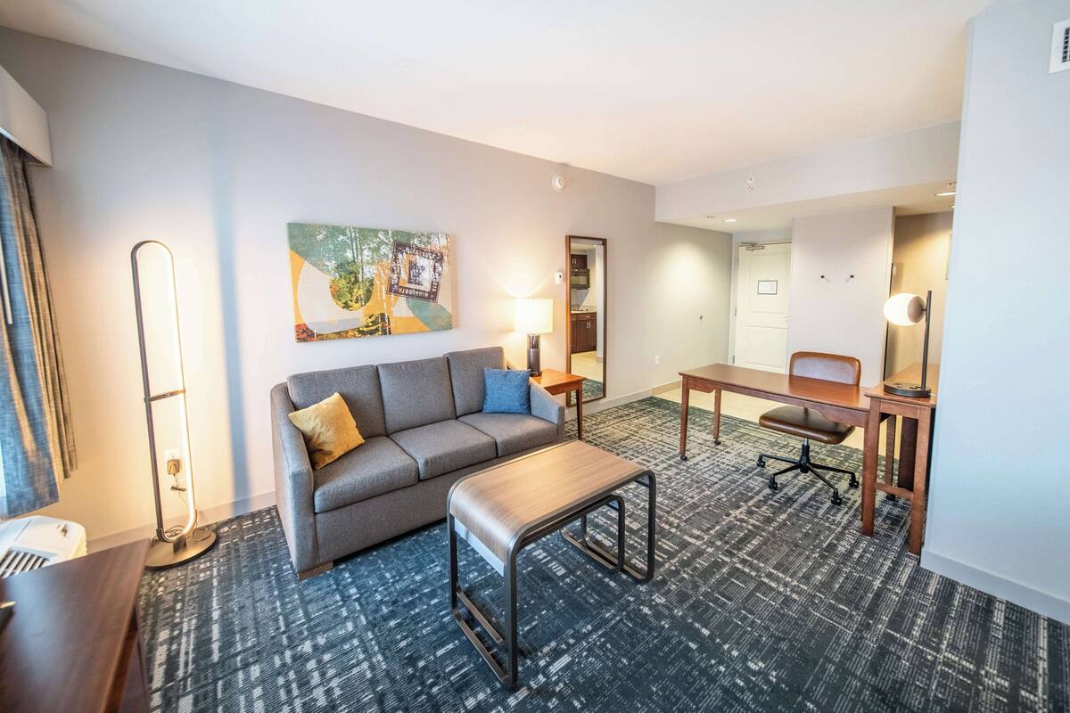 Een hotelkamer met een grijze bank, een kleine tafel, een bureau met een stoel, een lamp en een schilderij aan de muur.