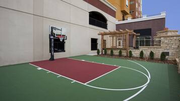 Una cancha de baloncesto al aire libre con superficie roja y verde, un aro de baloncesto y un muro de piedra con una pérgola de madera.