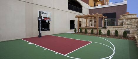 An outdoor basketball court with a red and green surface, a basketball hoop, and a stone wall with a wooden pergola.