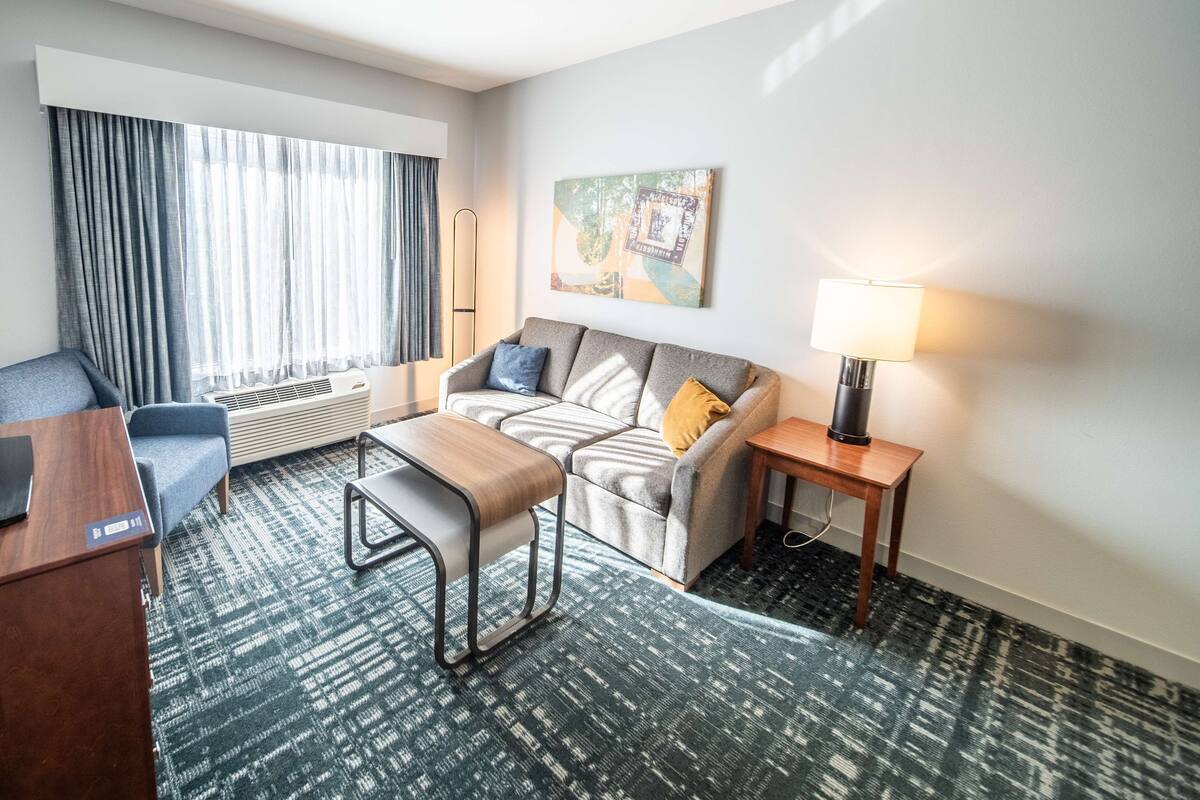 Een hotelkamer met een bank, een kleine tafel, een lamp, een stoel, een bureau en een groot raam met gordijnen.
