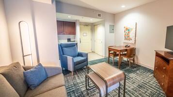 A hotel room with a sofa, a chair, a small table, a TV, a kitchen area, and a bathroom.