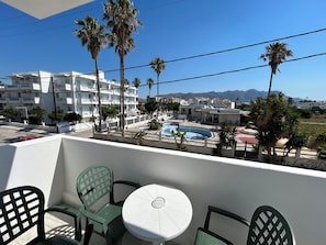 A balcony with two green chairs and a white table, overlooking a pool area with palm trees and a mountain in the background. - Kaloxenia Apartments (Kos)