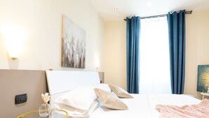 A hotel room with a bed, a bedside table with a vase, a painting on the wall, and blue curtains. - Cesar Vatican Rooms (Rome)