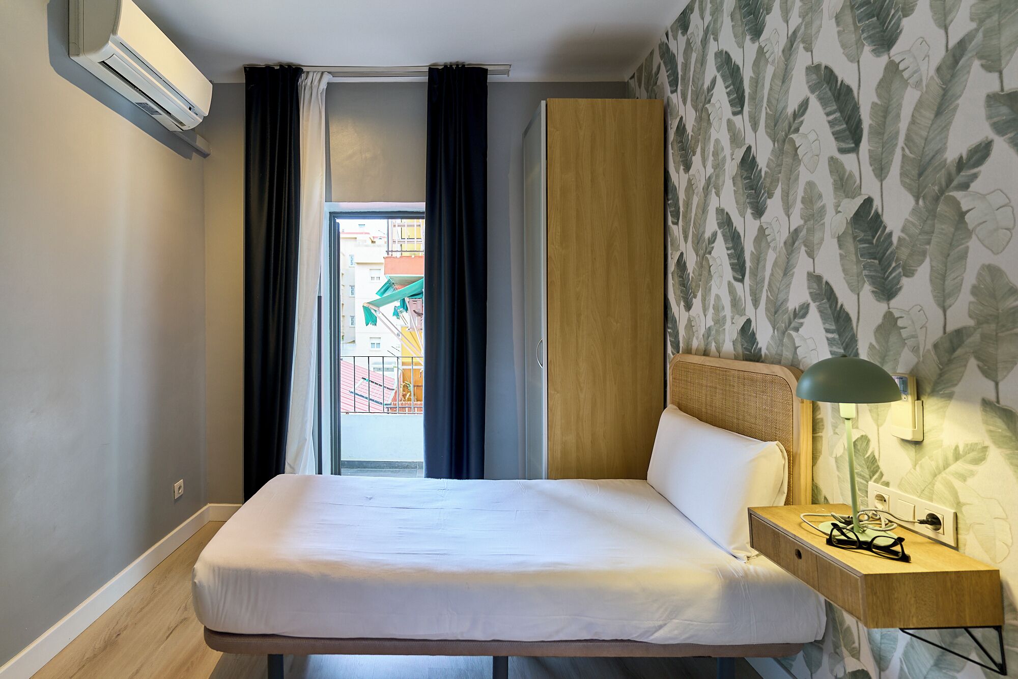 Una habitación de hotel con una cama, una mesita de noche con lámpara, una ventana con cortinas y una pared decorada con un diseño de hojas.
